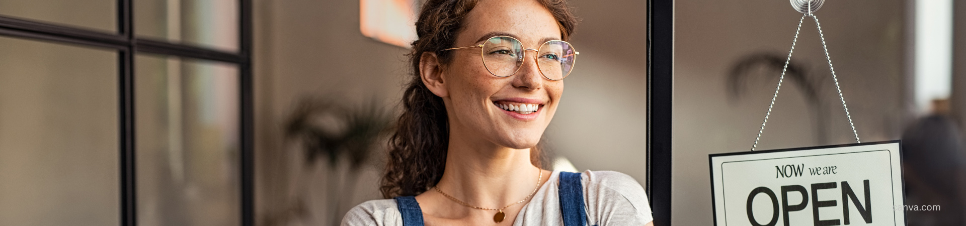 young woman standing in front of store with sign Now we are OPEN photo: canva.com